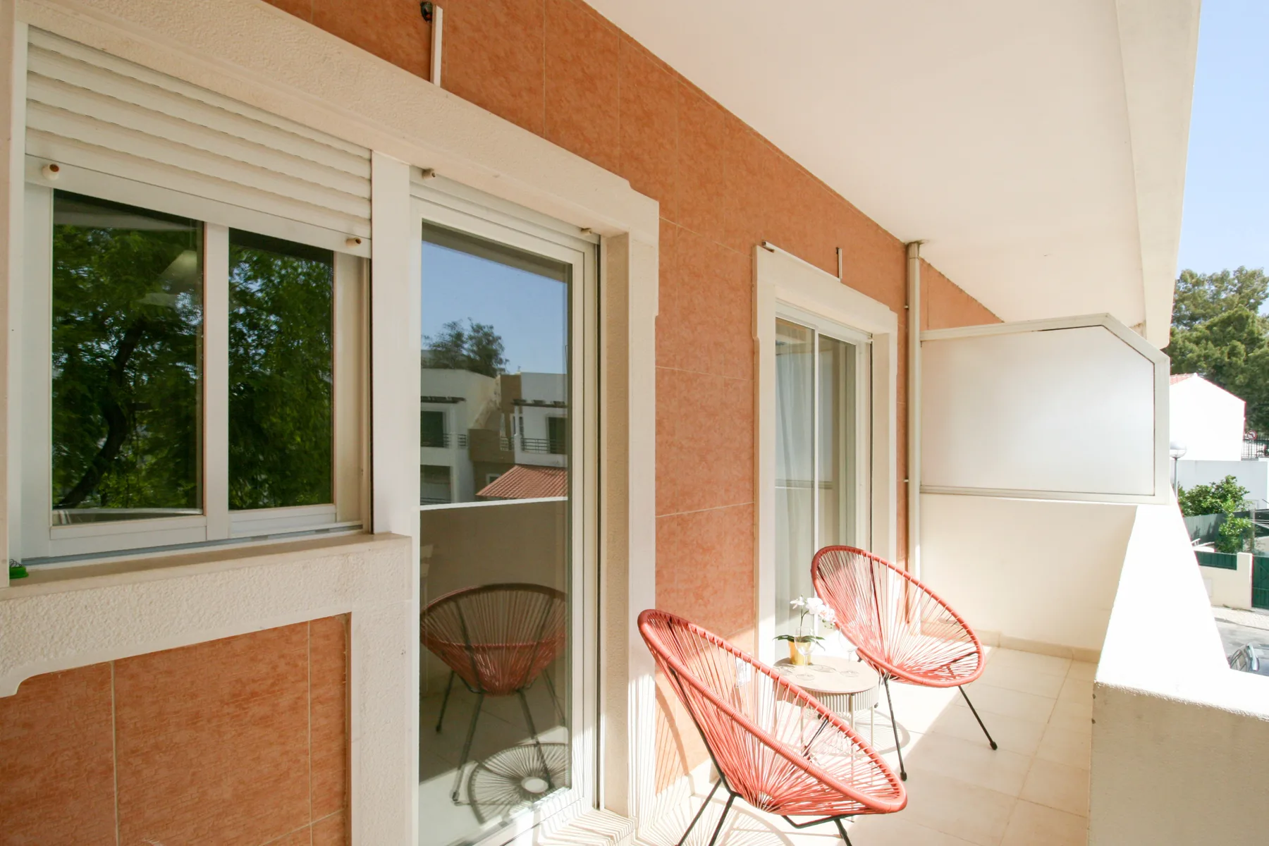 Un balcón con azulejos tiene puertas correderas de vidrio y asientos rojos de alambre.