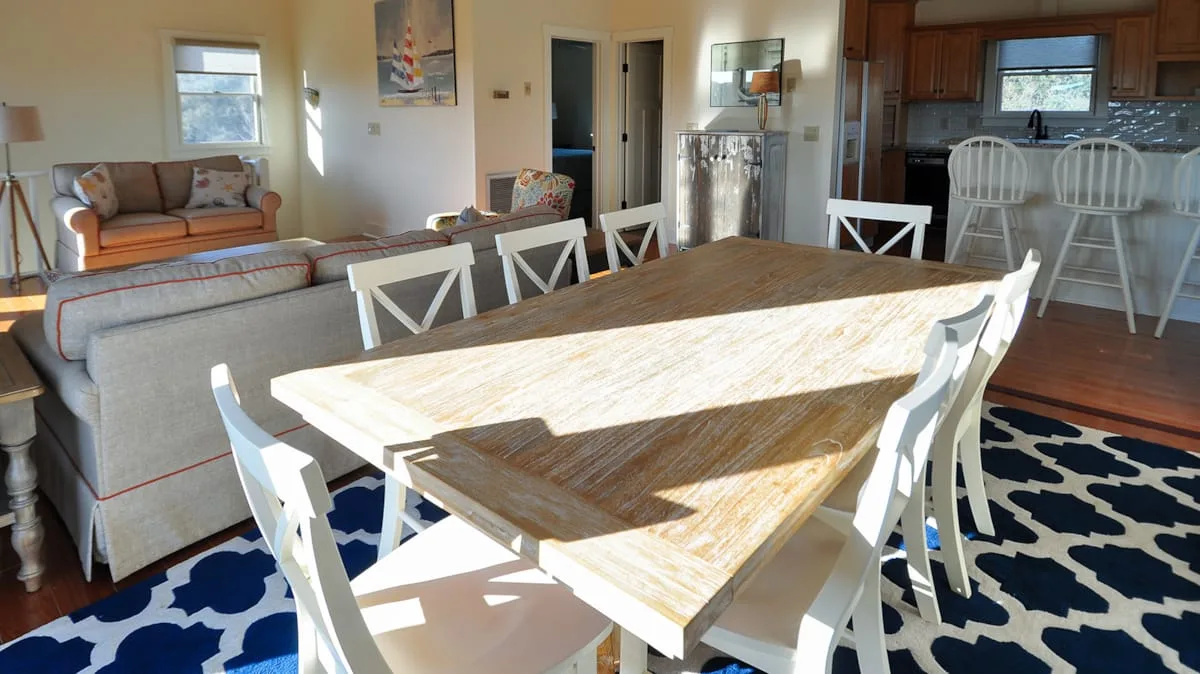 Comedor con una larga mesa de madera y sillas blancas junto a una sala de estar.