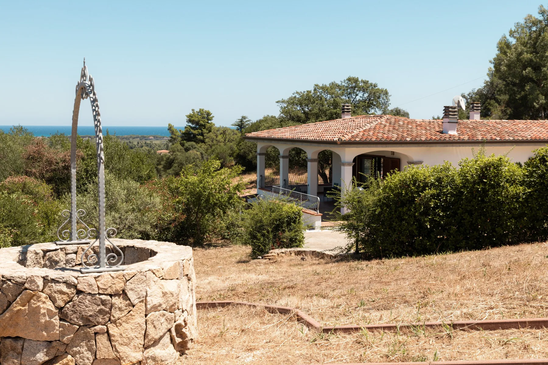 A house with a tiled roof sits among dry grass and shrubs, with the sea visible beyond.