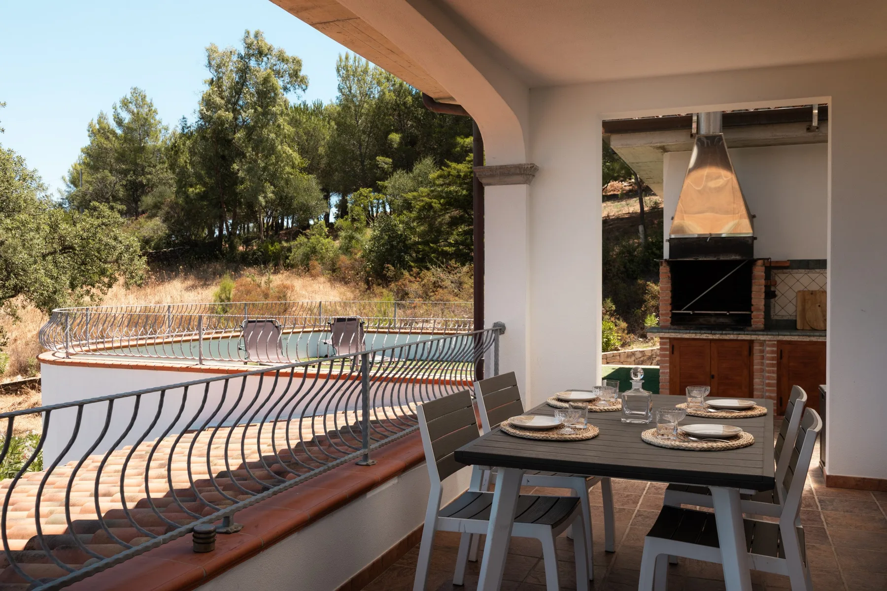A covered patio dining area overlooks a swimming pool and wooded hillside.