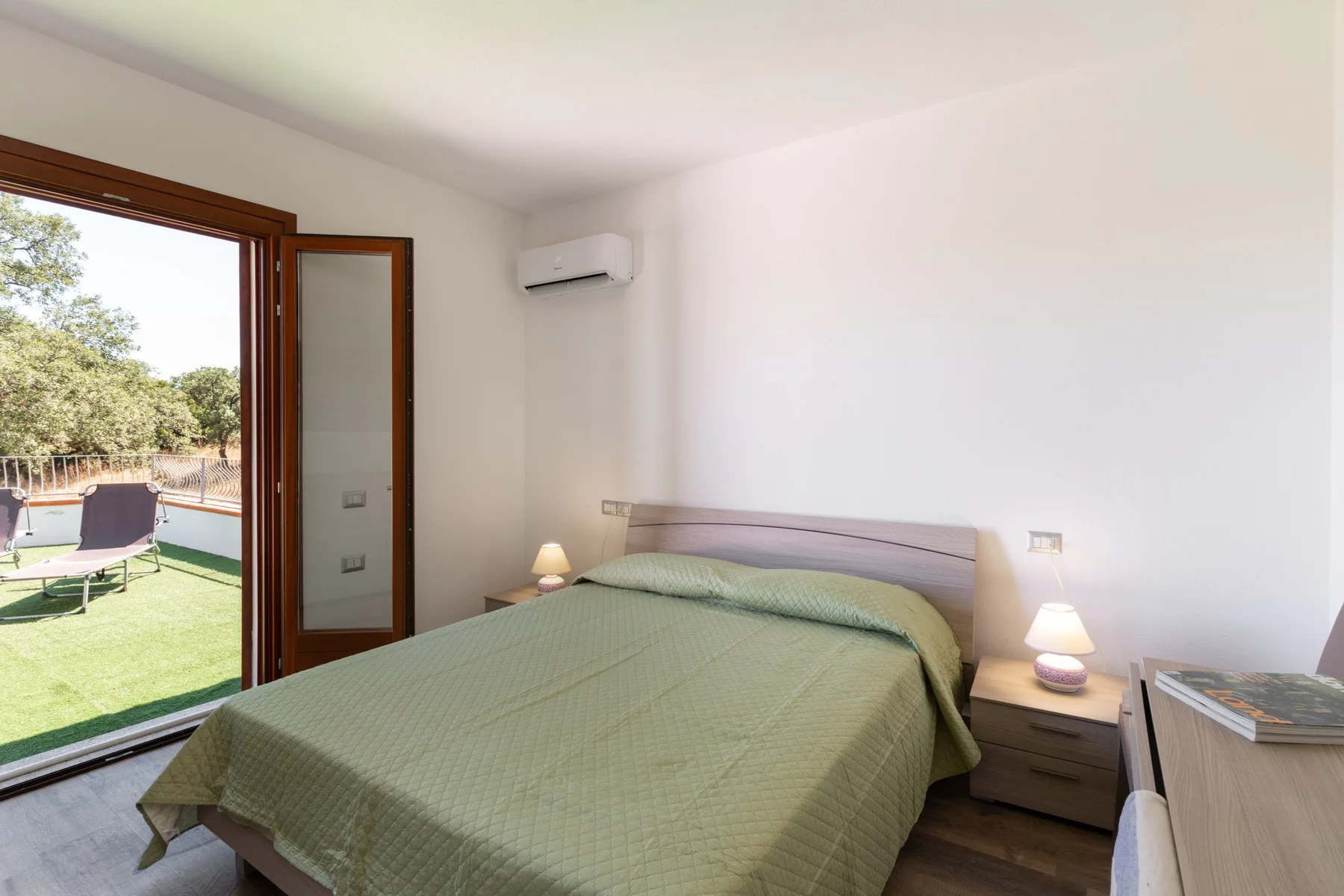 A bedroom with a bed beside an open doorway to a green outdoor area.