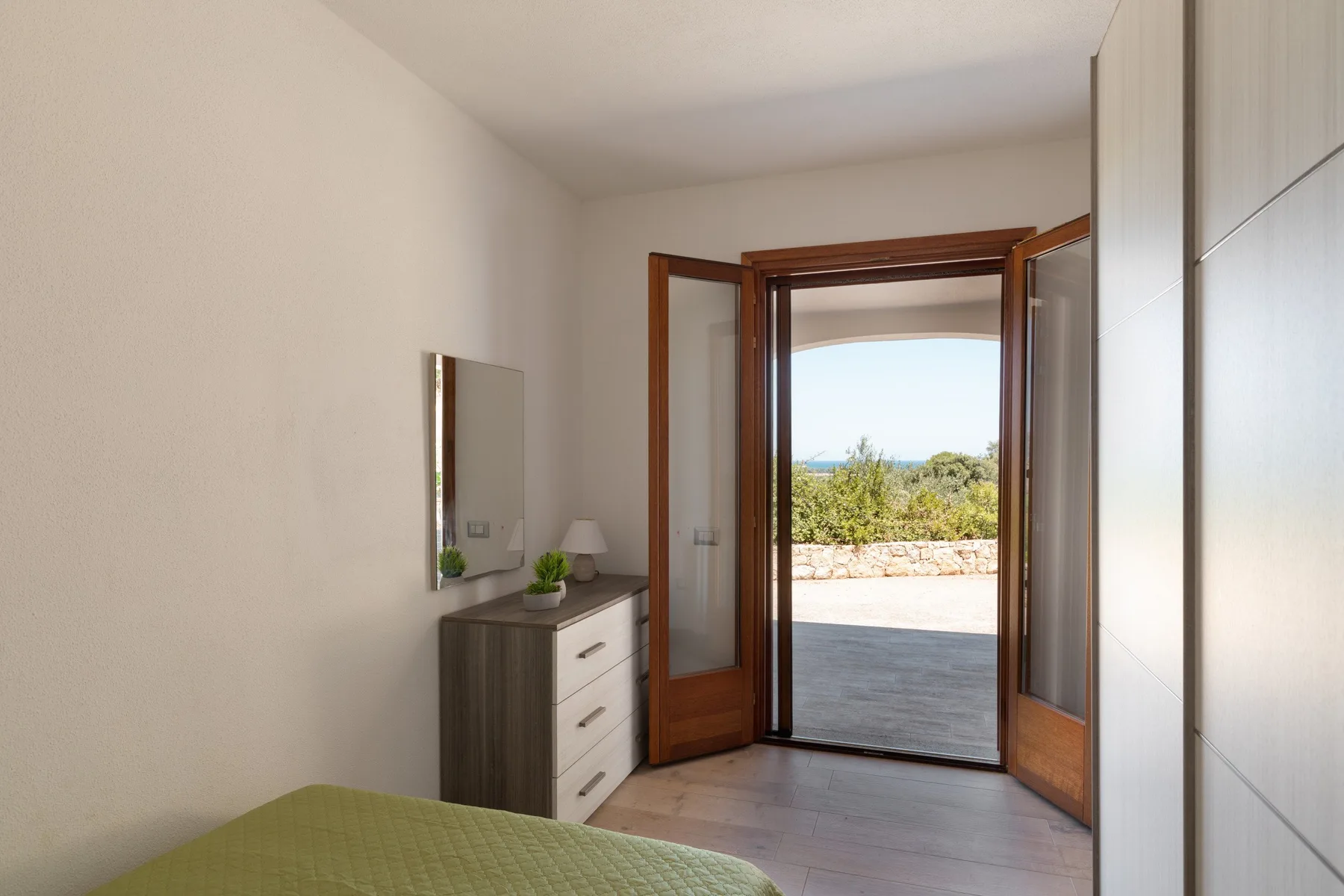 A bedroom opens through glass doors onto a sunlit terrace with greenery beyond.