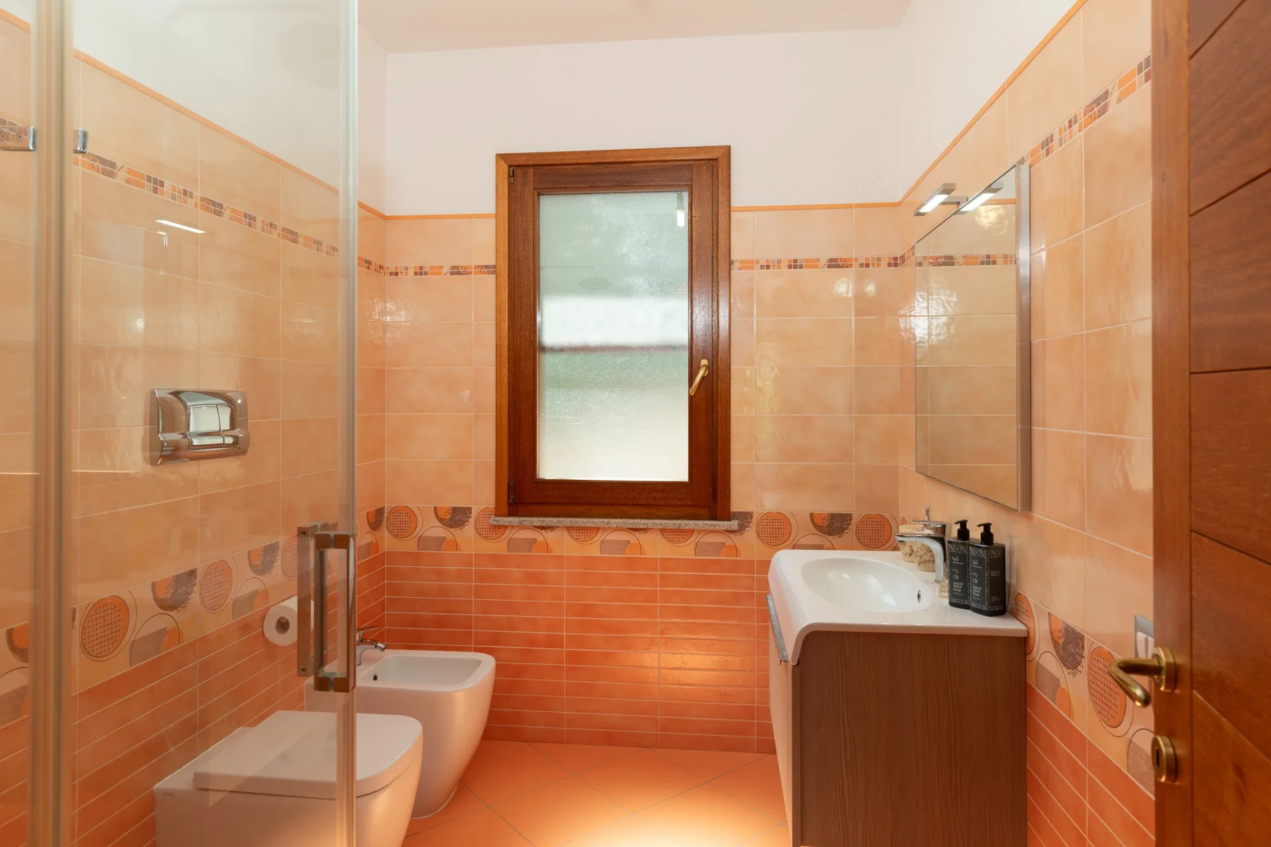 A tiled bathroom with a glass shower, toilet, and sink beneath a window.