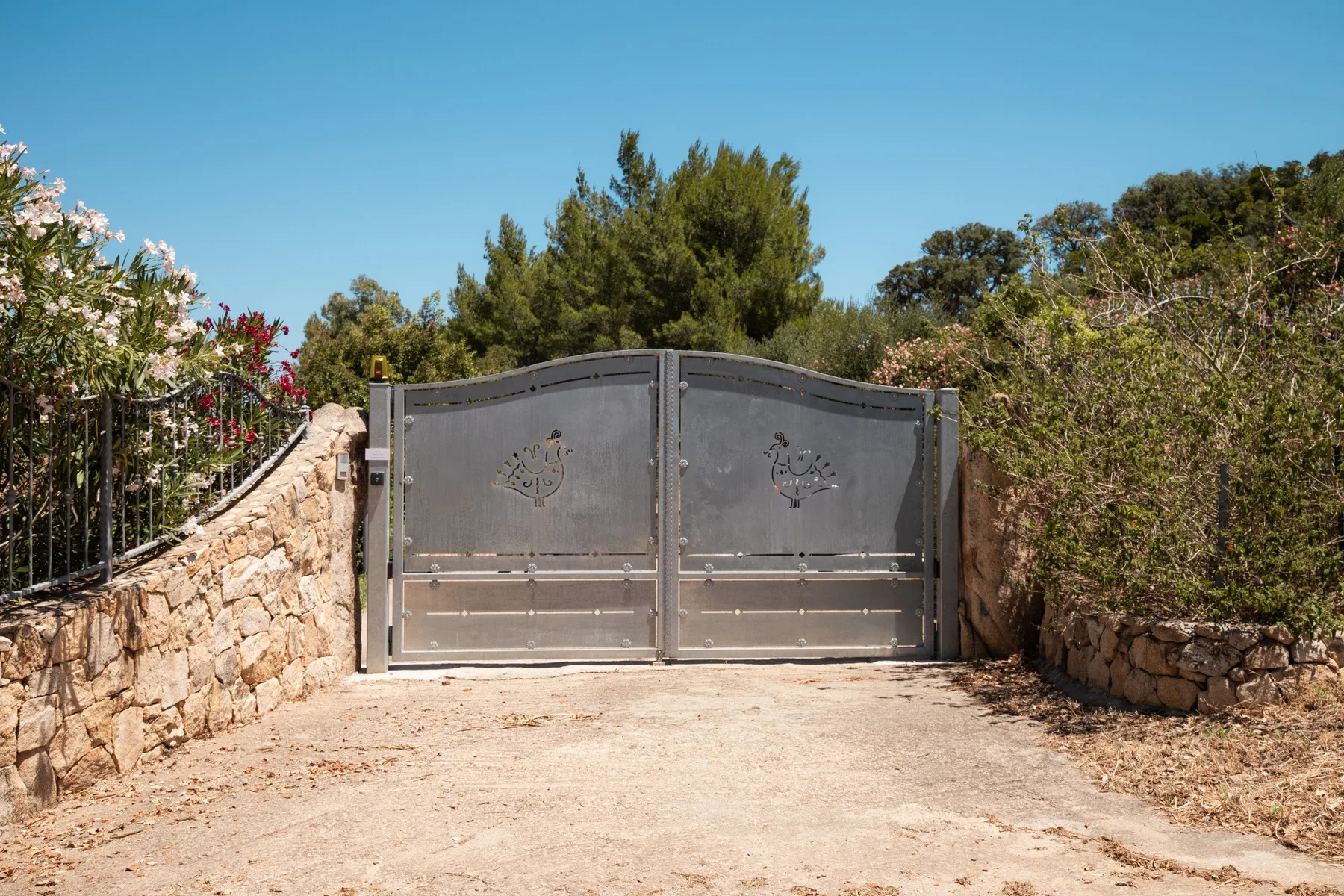 A closed metal gate stands at the end of a gravel driveway, bordered by stone walls and shrubs.