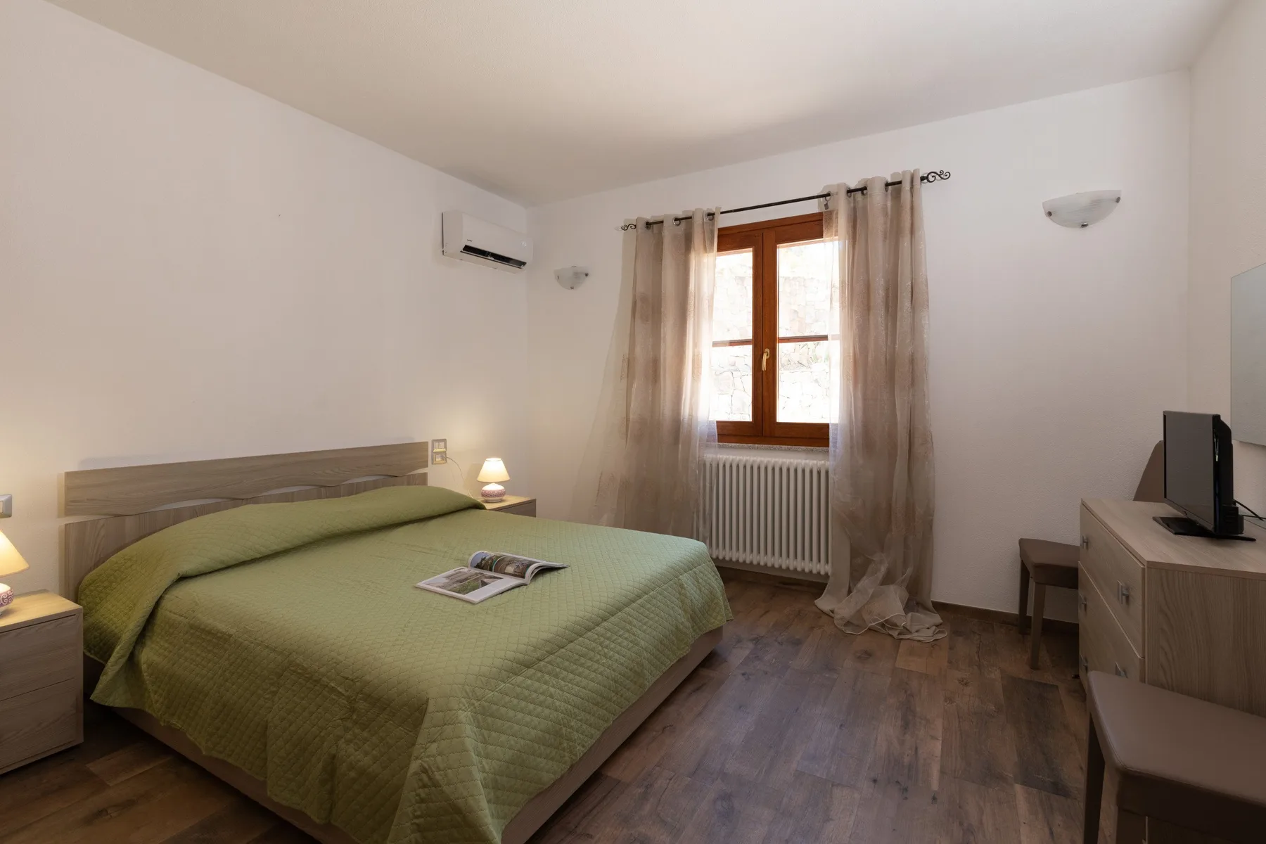 A bedroom with a green-covered bed, a curtained window, and a television.