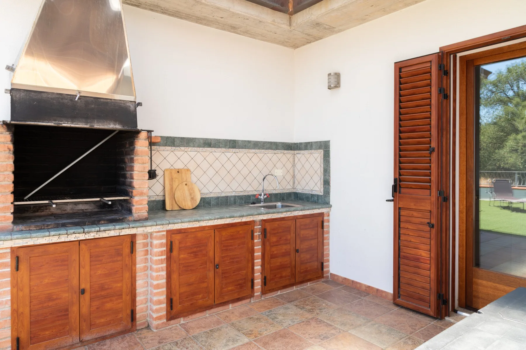 An outdoor cooking area with a brick grill, counter, and sink beside an open doorway.