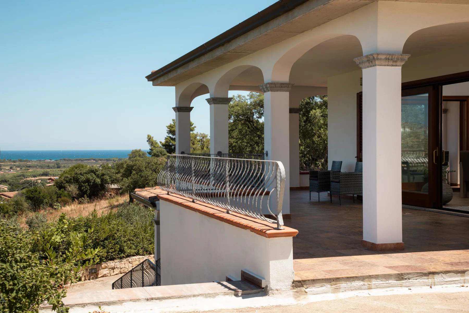 A covered patio with arched columns overlooks a green landscape and distant sea.