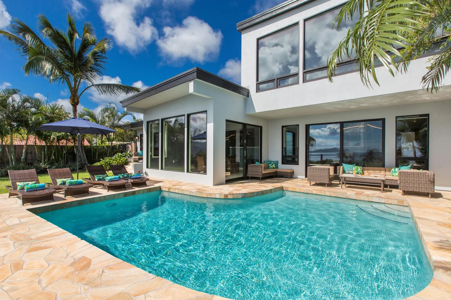 Piscina exterior junto a una casa moderna, con asientos en el patio y palmeras.