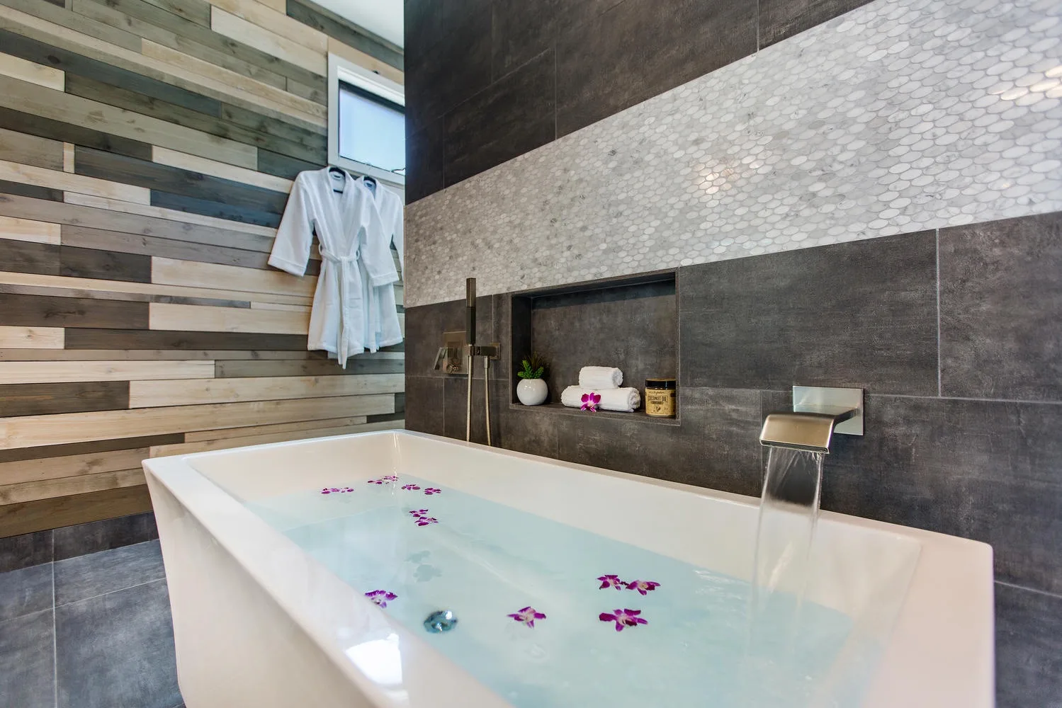 Un baño cuenta con una bañera exenta junto a una pared de azulejos oscuros.