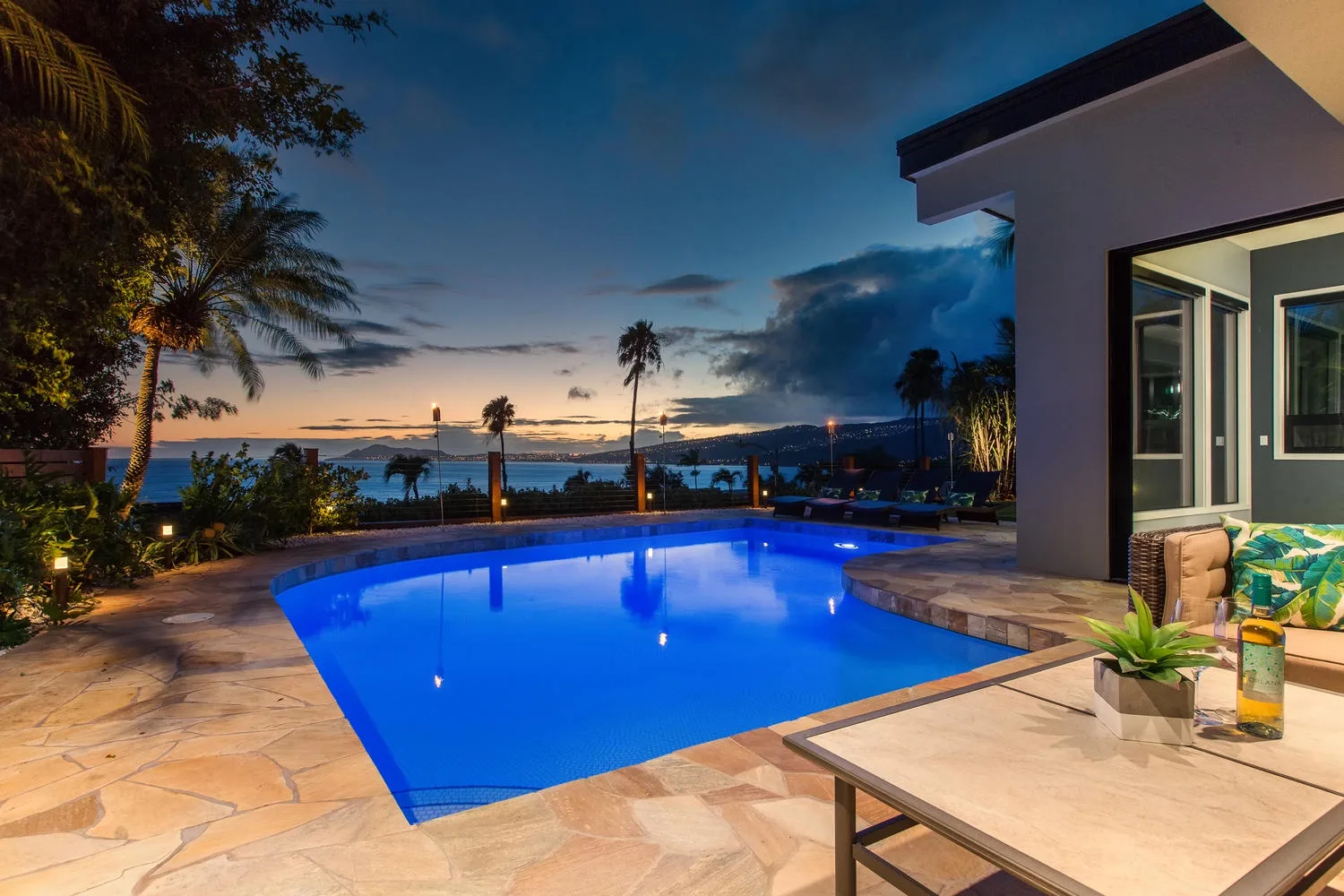 Una piscina iluminada está junto a una casa moderna al atardecer, con vistas a la costa.