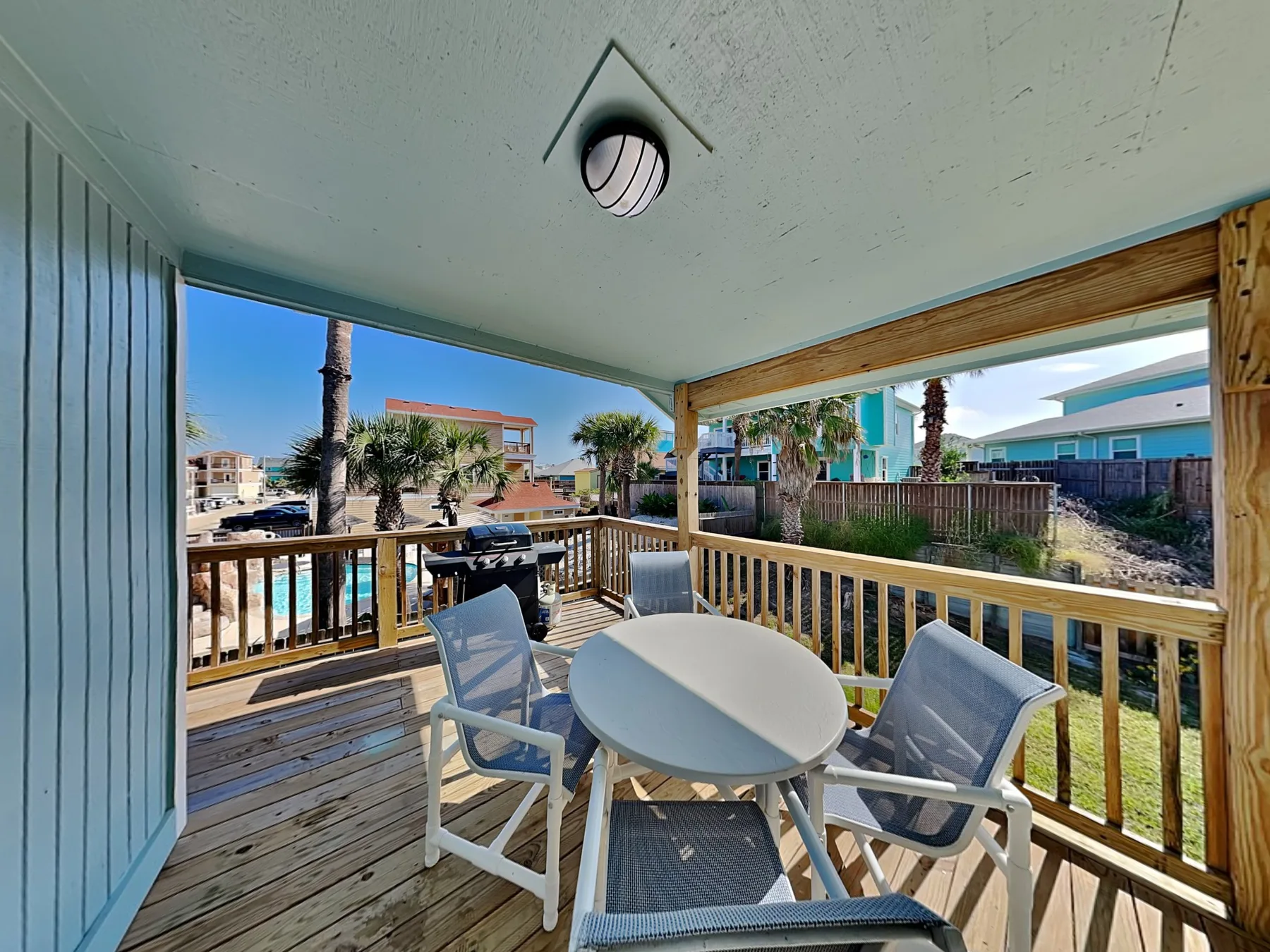 A covered outdoor deck has a round table and seating overlooking nearby homes.