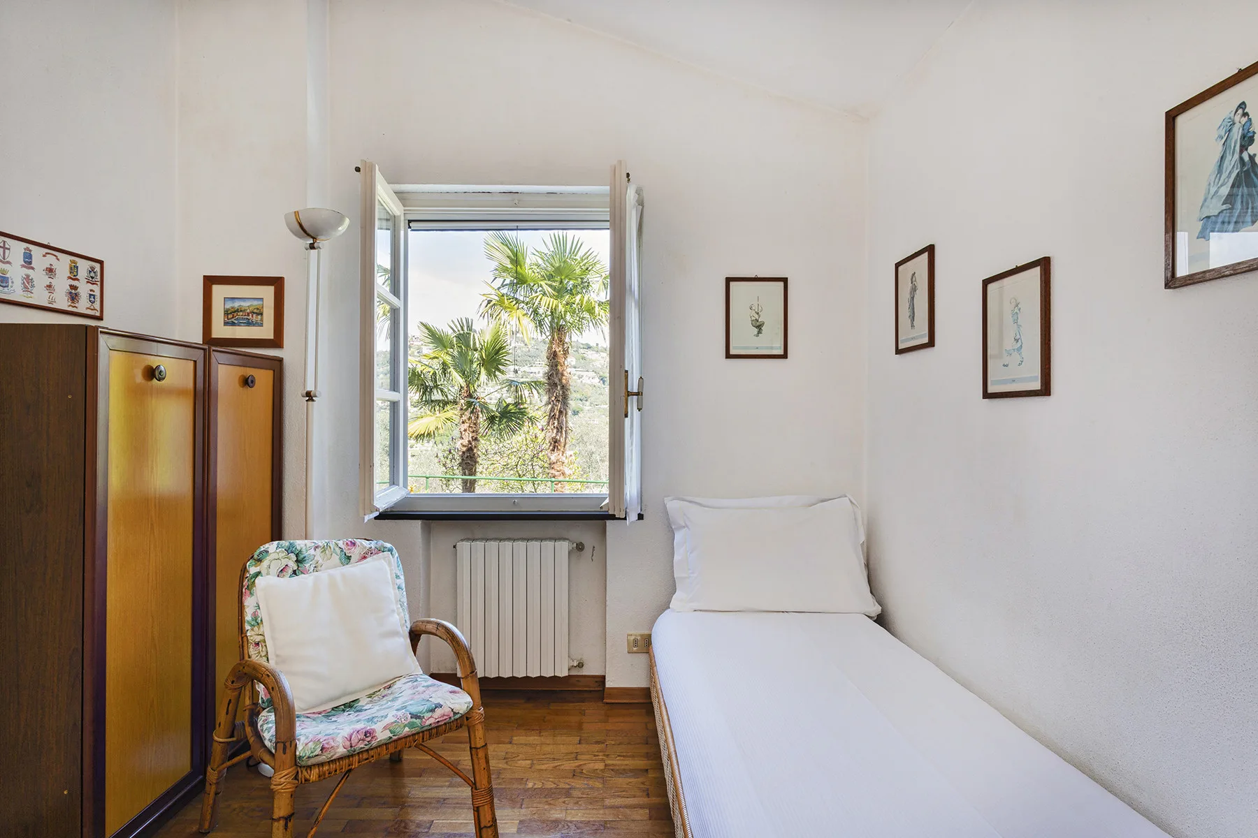 Un dormitorio con una cama individual junto a una ventana abierta con palmeras a la vista.