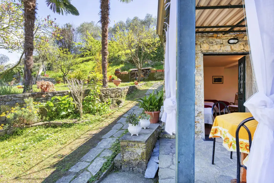Un sendero de jardín junto a un patio de piedra, con mesa y sillas bajo una zona cubierta.