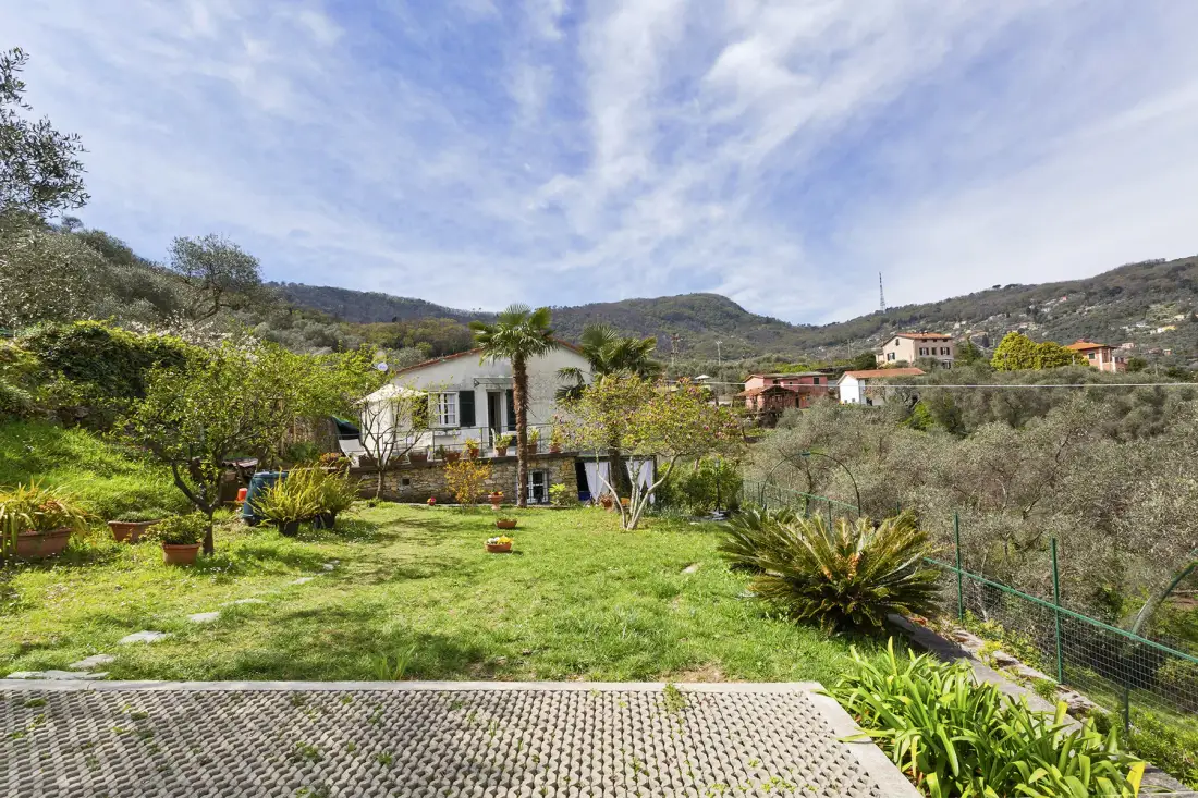 Una casa está junto a un jardín con césped, con colinas y casas dispersas al fondo.