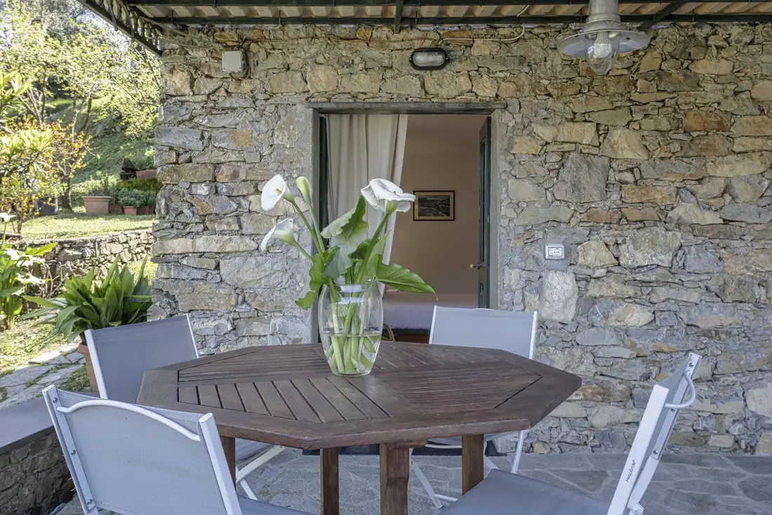 Un comedor en un patio de piedra tiene una mesa redonda, asientos blancos y un florero con flores.