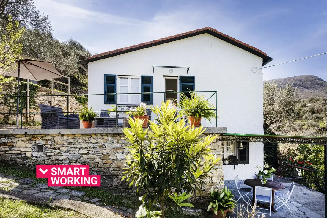 Una casa blanca con terraza y asientos al aire libre se alza sobre un muro de piedra.