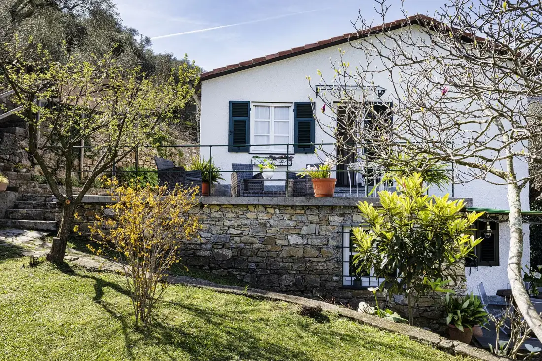Una casa blanca se alza sobre un muro de piedra, con un jardín delante.
