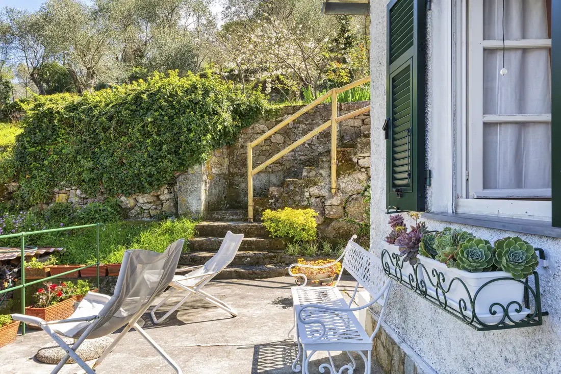 Un patio soleado tiene asientos junto a un jardín y una ventana con contraventanas.