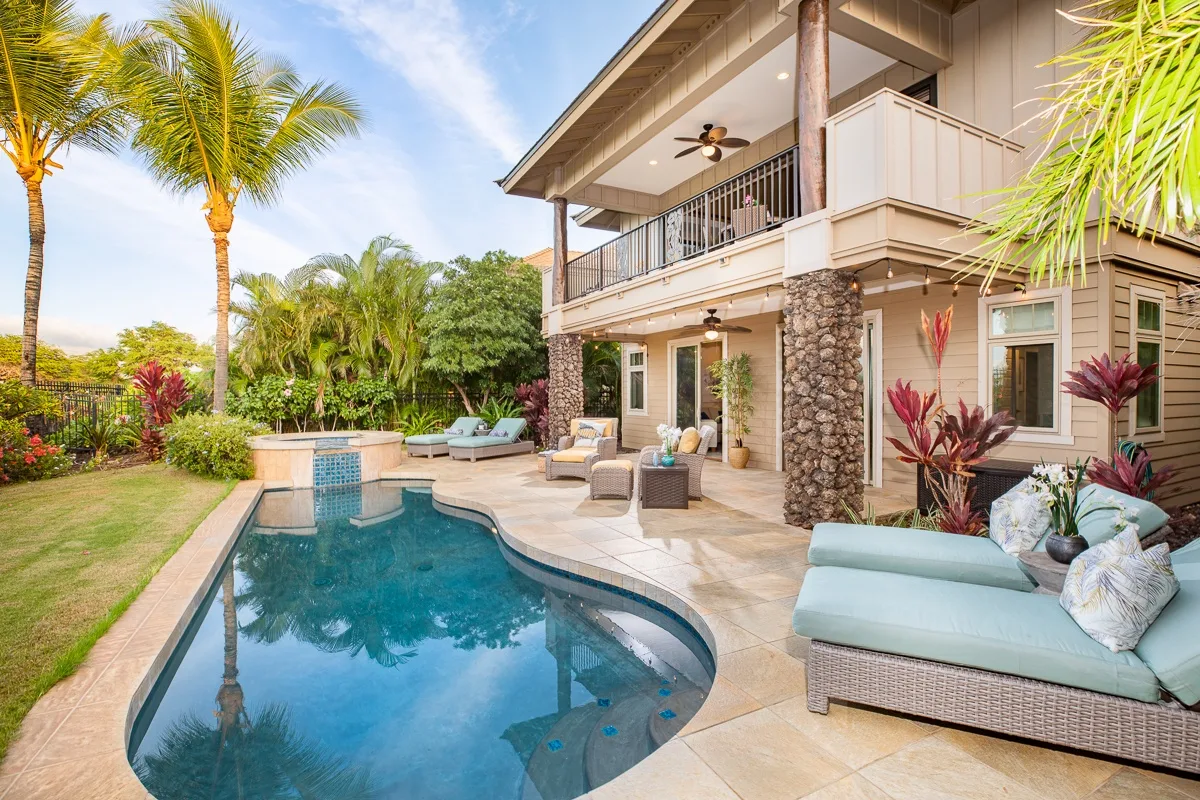 Una piscina tropical al aire libre junto a una casa de dos pisos y asientos en el patio.