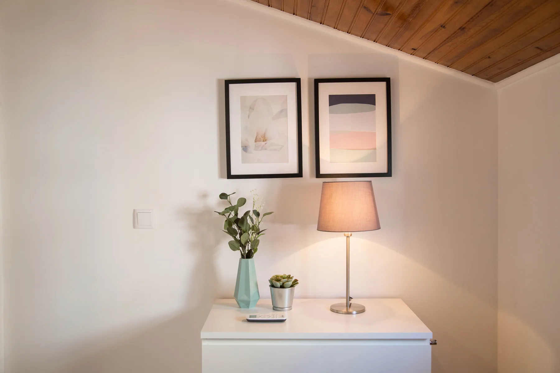 A small table with a lamp, framed artwork, and plants sits against a white wall.