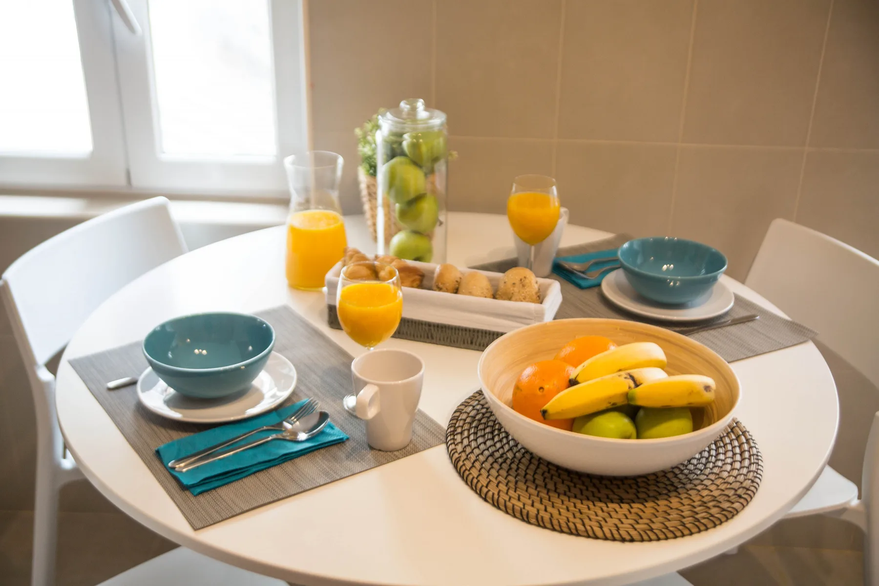 A dining table is set with bowls, fruit, and breakfast items beside a window.