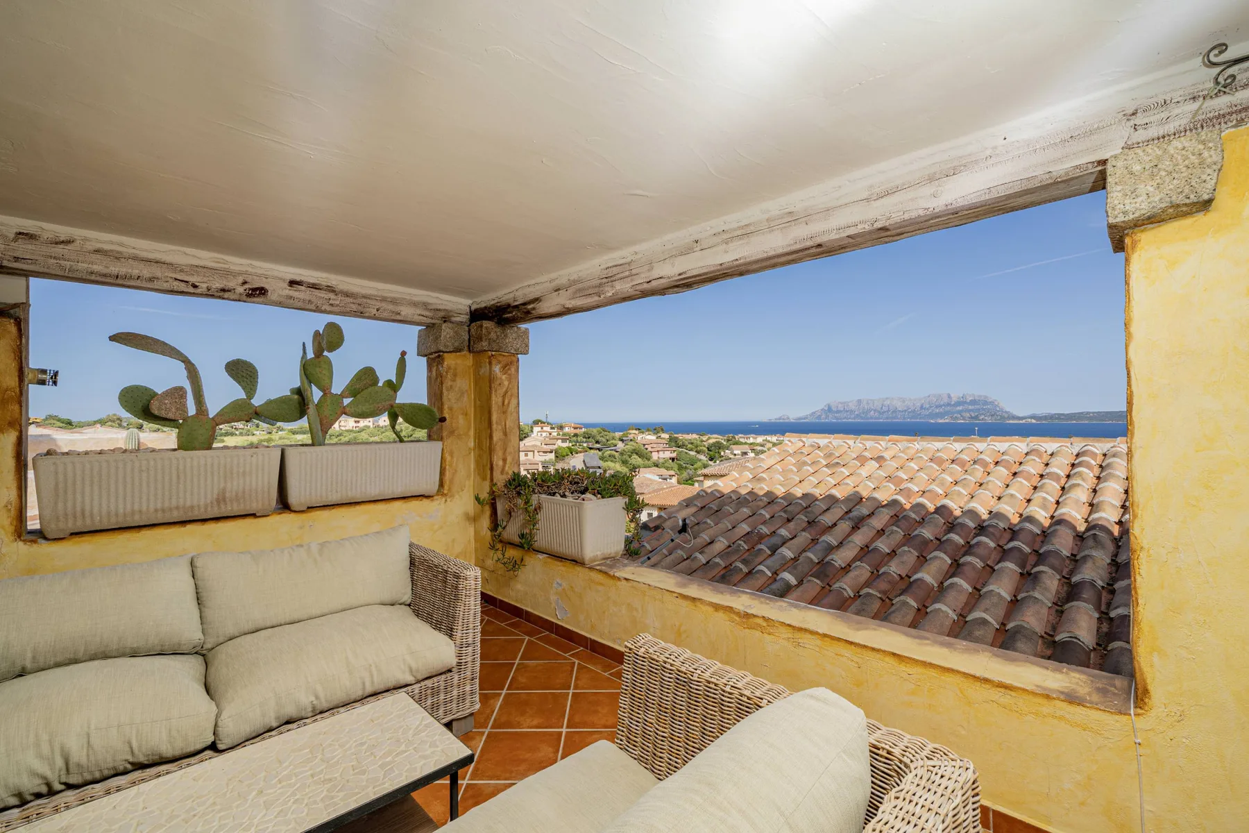 Una terraza cubierta tiene asientos acolchados y vistas a los tejados y al mar.