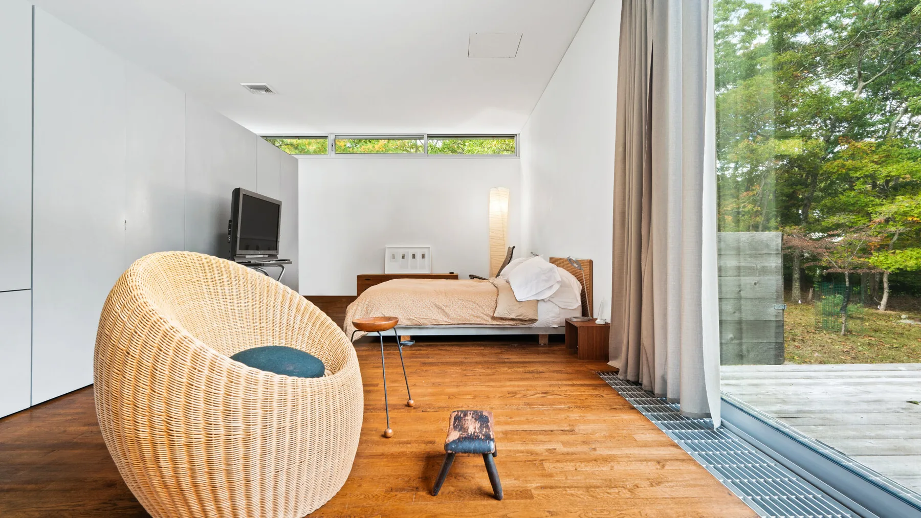 A bright bedroom with a bed, woven seating, and large windows overlooking greenery.