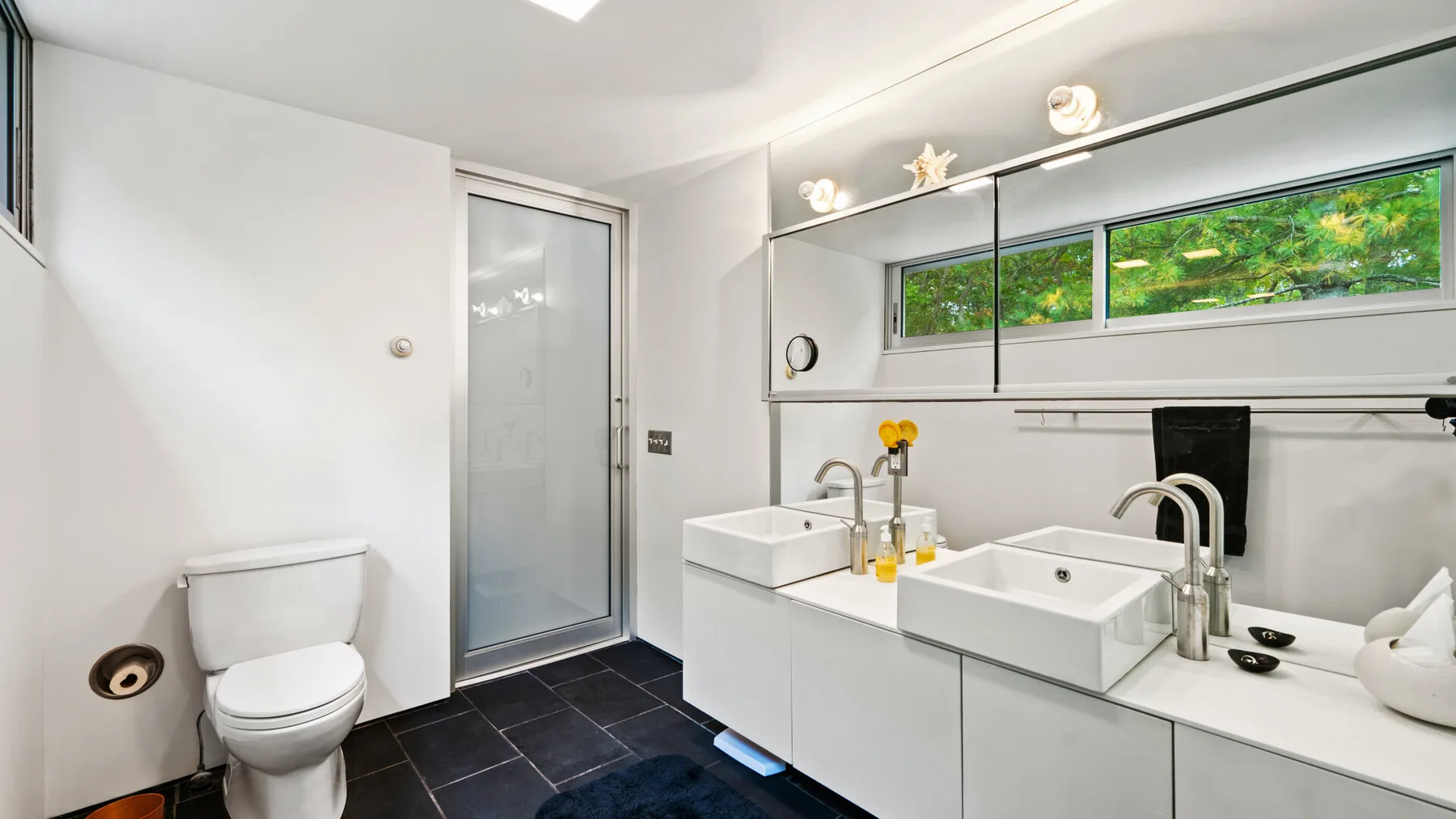 A bright bathroom features a long counter with two sinks and a toilet.