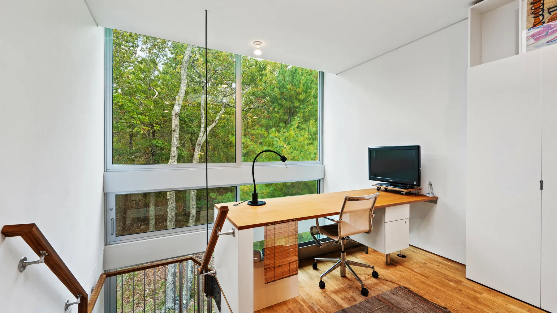 A bright home office with a desk, computer, and large windows overlooking trees.