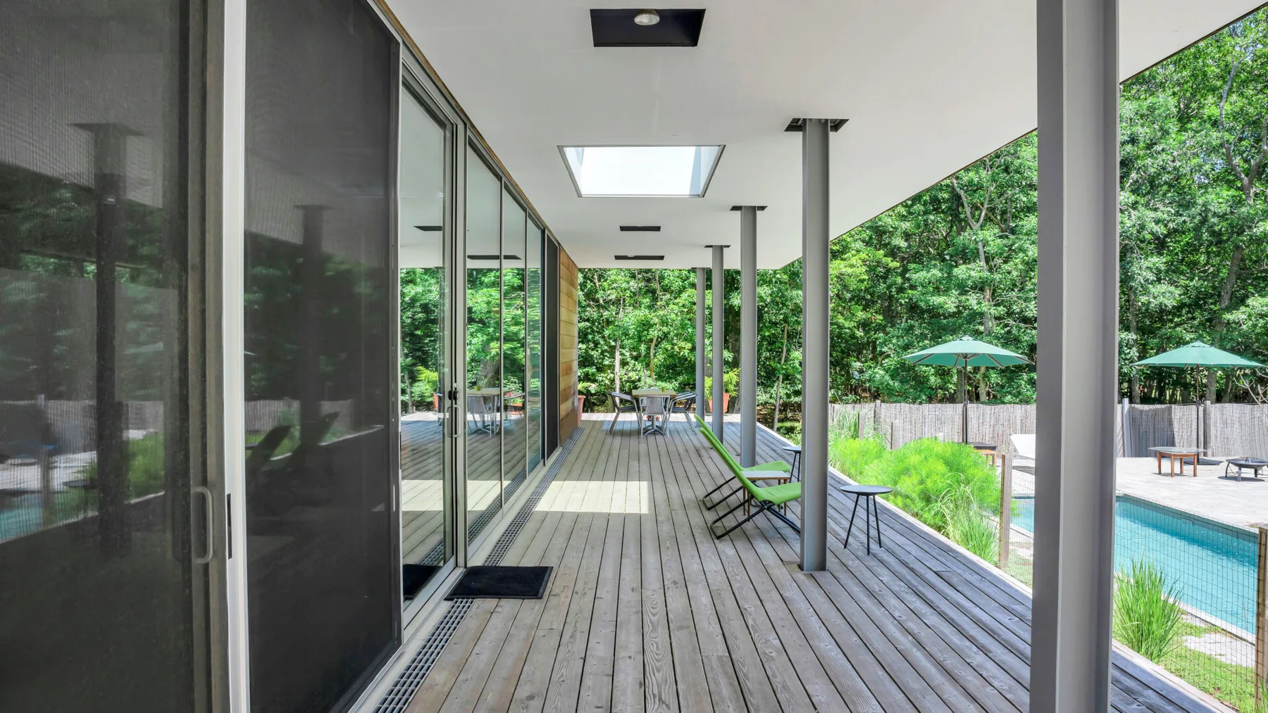 A covered outdoor deck with glass doors overlooks a swimming pool and trees.