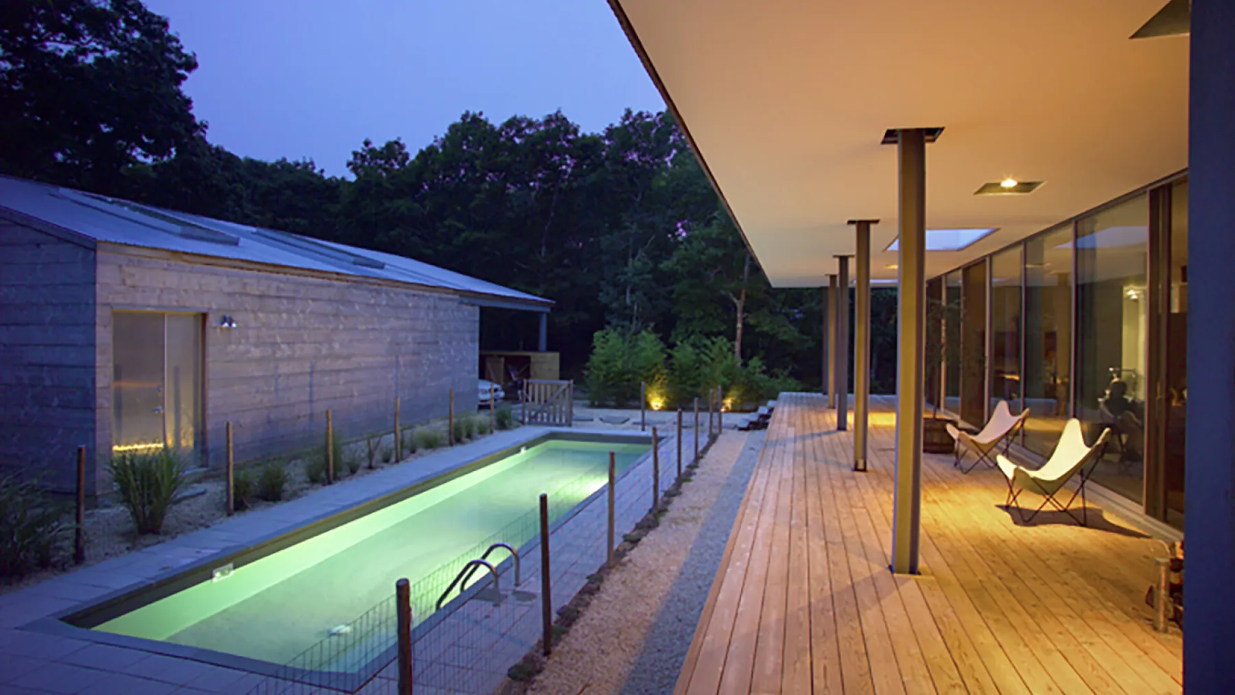 A modern outdoor pool beside a wooden deck and glass-walled building at dusk.