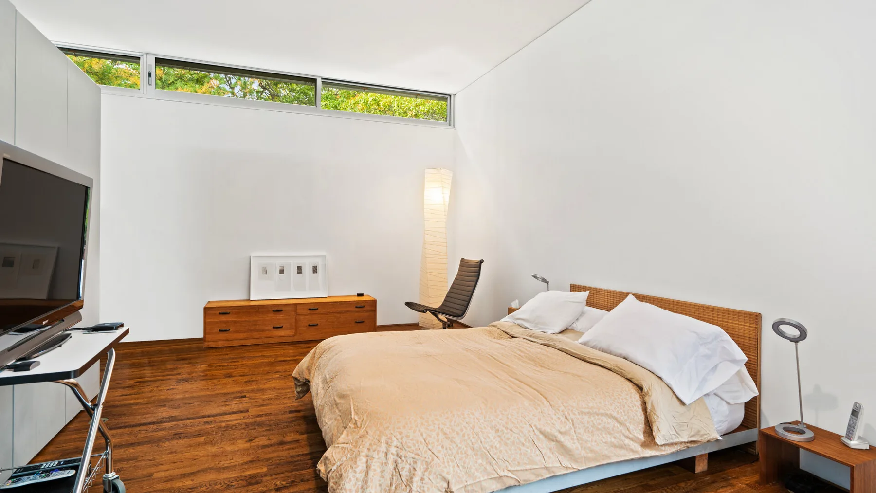 A bright bedroom with a bed, wooden dresser, and high windows.