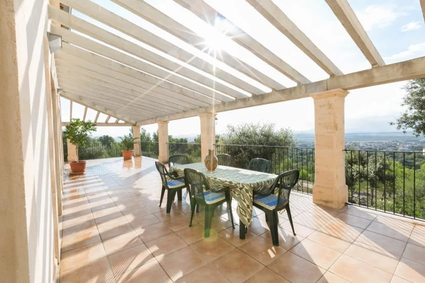 A sunlit outdoor dining area sits beneath a wooden pergola, overlooking the landscape.
