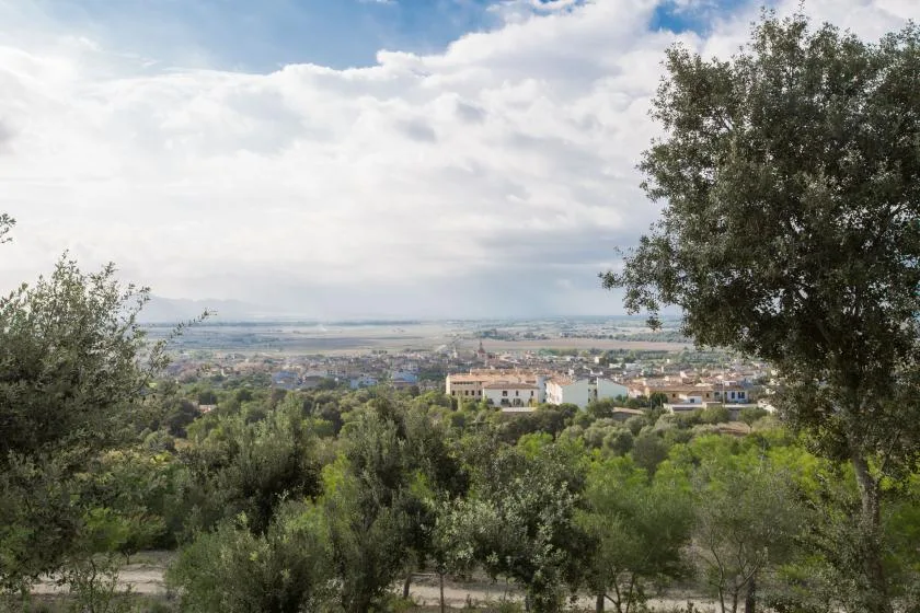 A hillside landscape overlooks a town and fields beneath a cloudy sky.