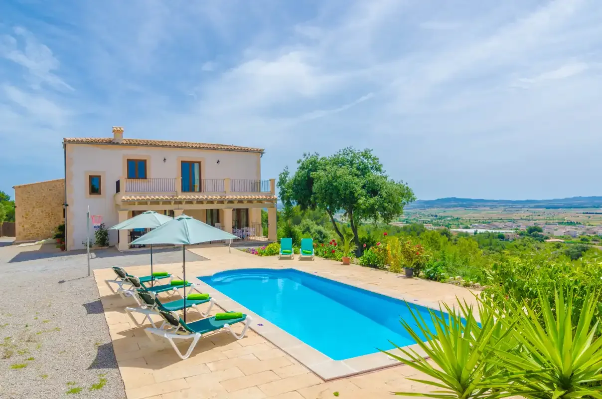 A house overlooks a swimming pool and lounge seating on a sunny terrace.