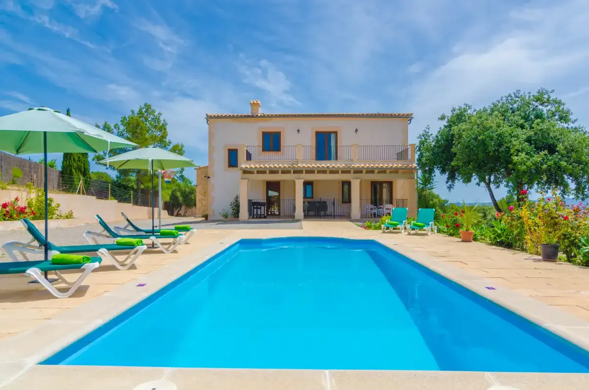 A swimming pool faces a two-story house, with lounge chairs and umbrellas along the patio.