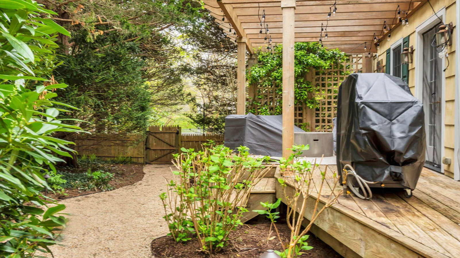 Un patio de jardín bajo una pérgola de madera, con un sendero de grava y plantas frondosas.