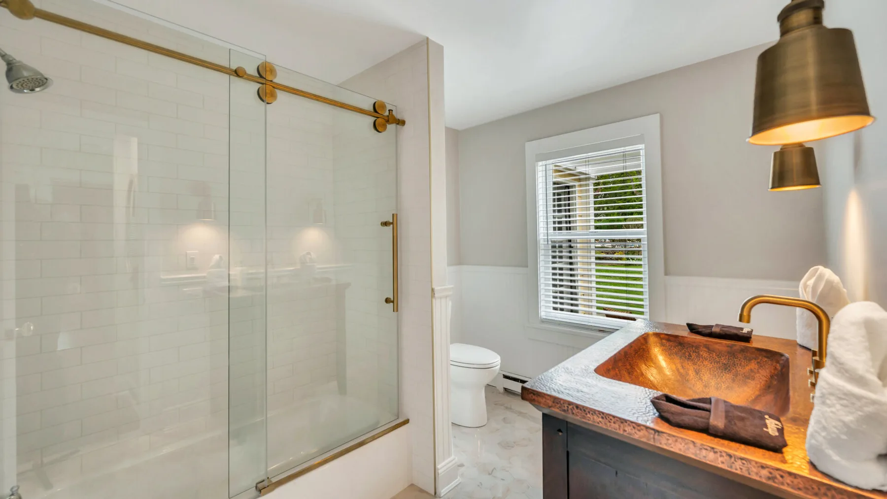 Un baño luminoso cuenta con una ducha de vidrio, un lavabo sobre la encimera y una ventana con persianas.