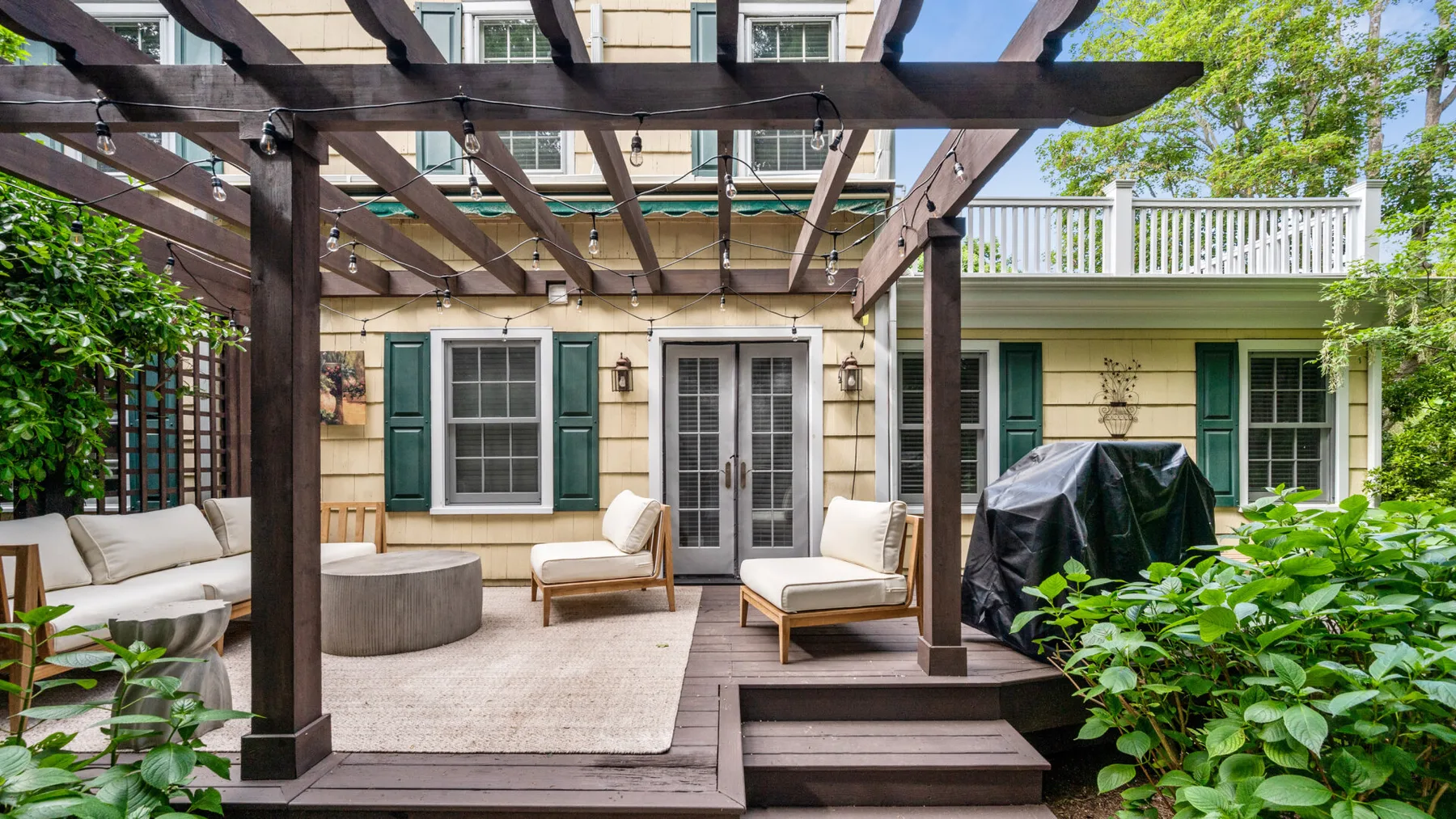 Un patio exterior amueblado bajo una pérgola de madera, junto a una casa.