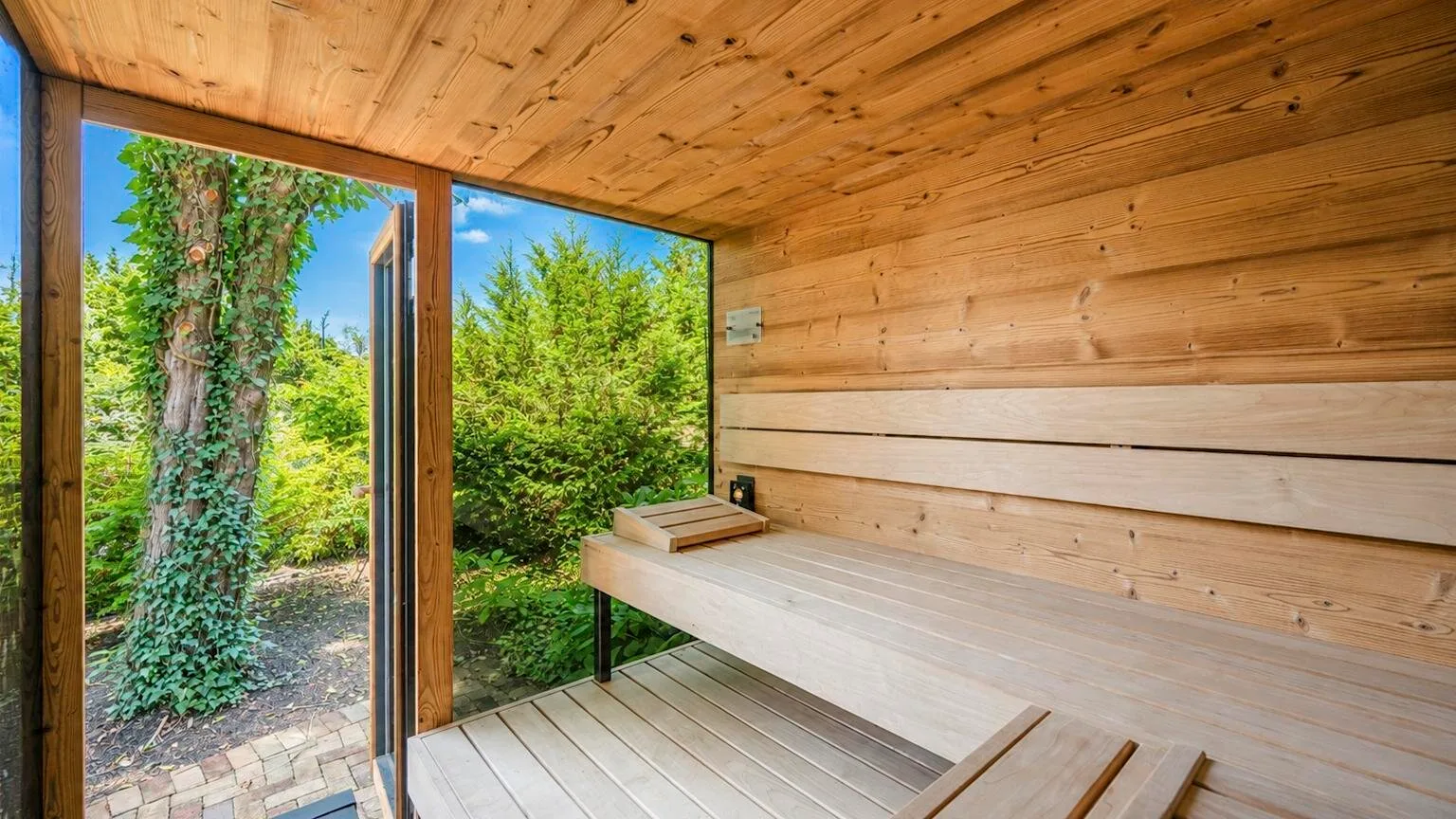 Interior de sauna de madera con un banco largo y una puerta de vidrio abierta hacia la vegetación.