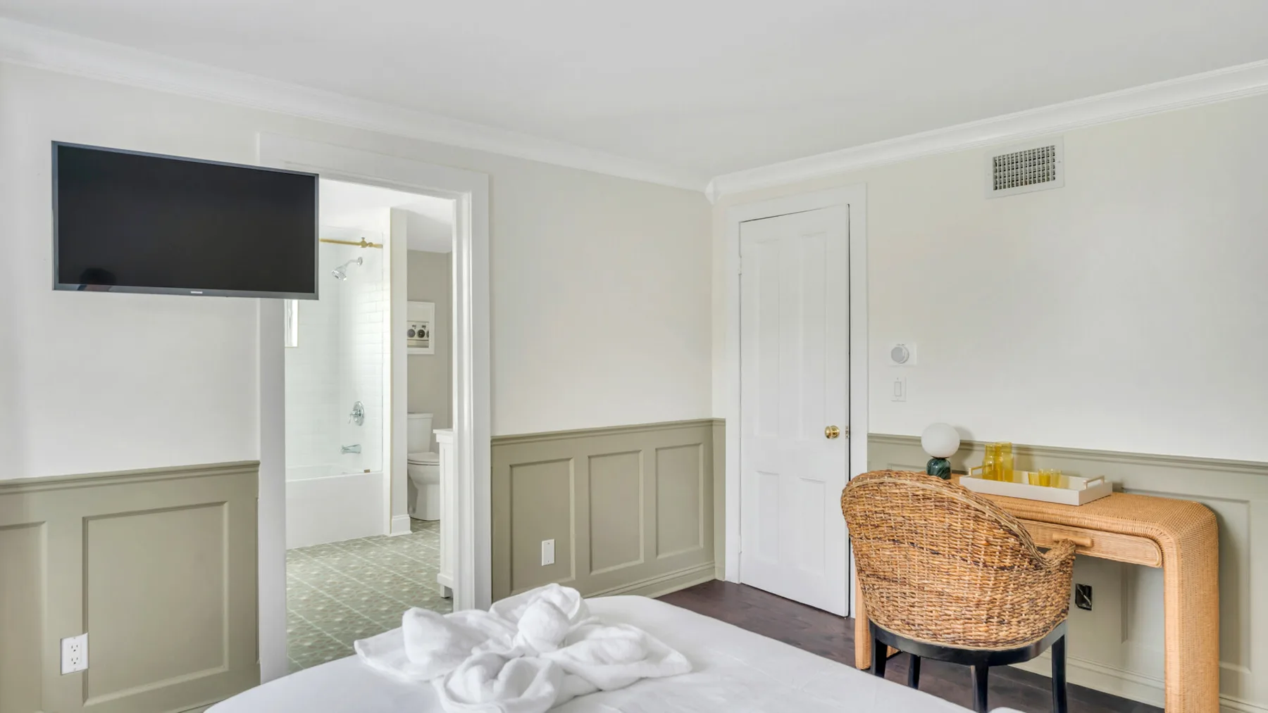 Un dormitorio con cama, televisor y silla tejida junto a una consola.
