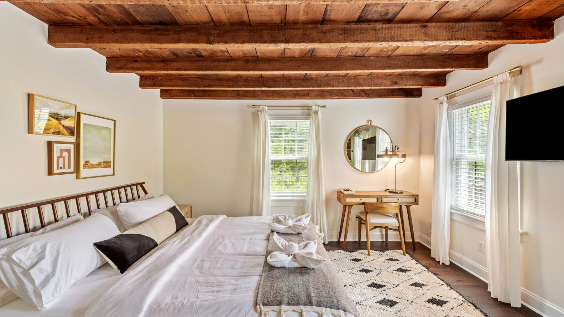 Un dormitorio con cama, escritorio pequeño y ventanas bajo vigas de madera expuestas.