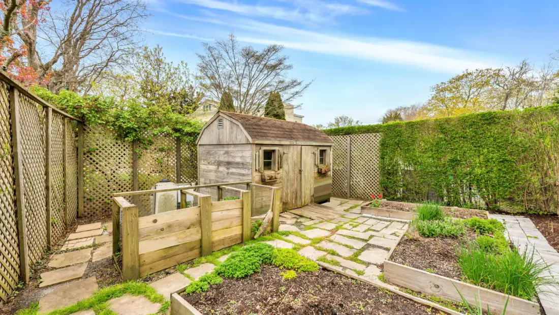 Un patio de jardín con una pequeña caseta de madera, bancales elevados y setos frondosos.