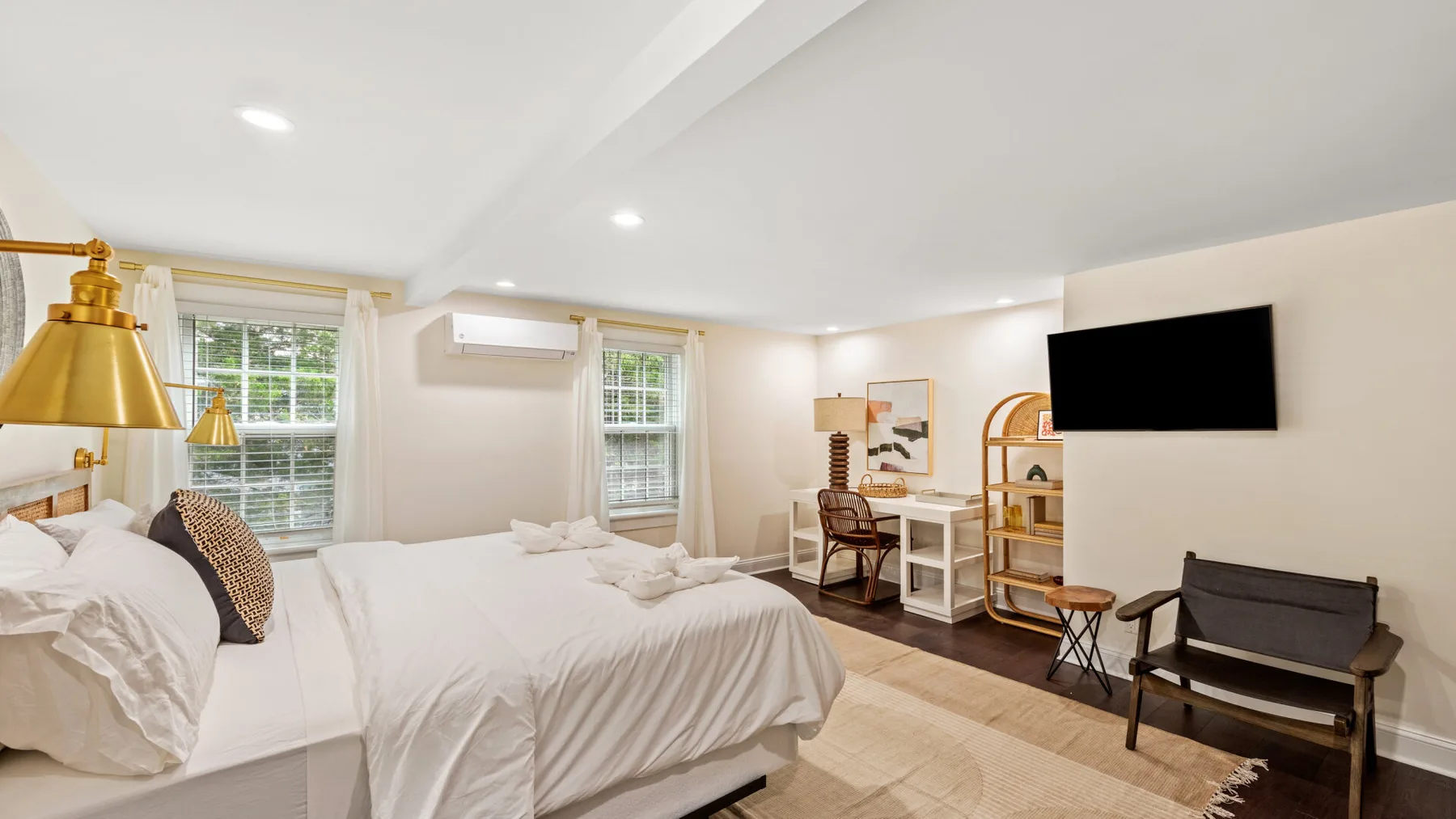 Un dormitorio luminoso con ropa de cama blanca, un televisor en la pared y asientos.