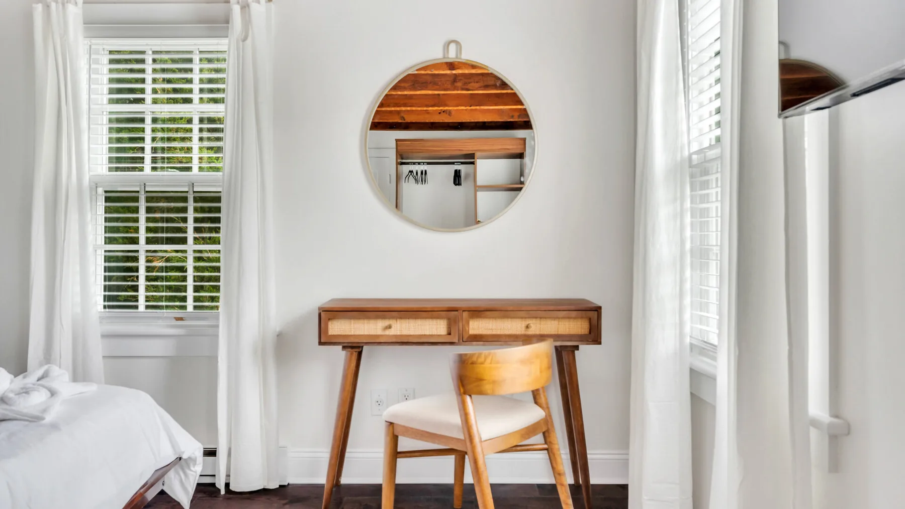 Dormitorio luminoso con escritorio, silla y espejo redondo entre dos ventanas.