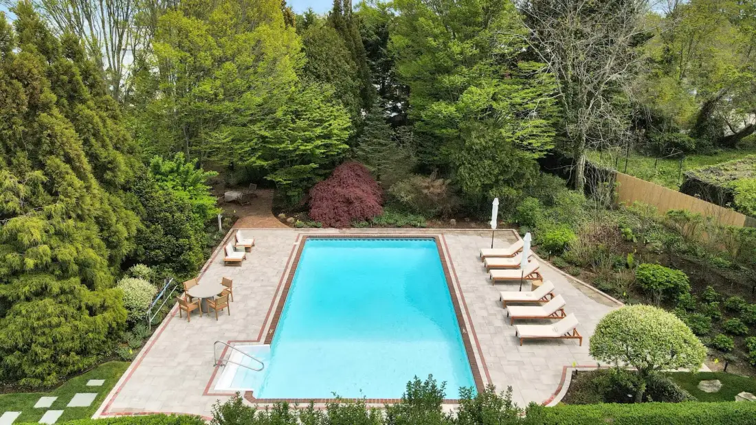 Una piscina rectangular está en una zona pavimentada del jardín, rodeada de árboles.