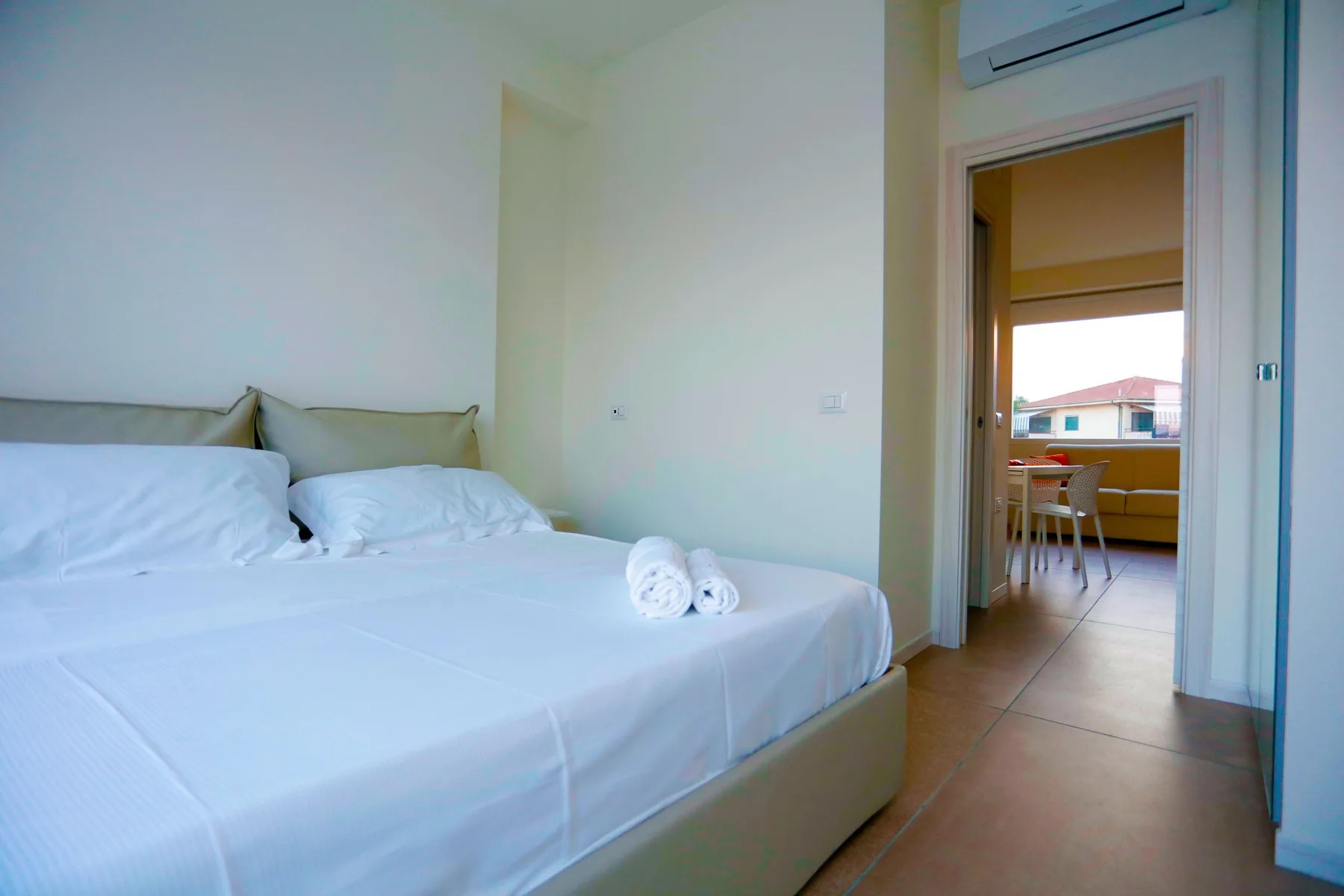 Un dormitorio luminoso con una cama bien tendida y vista a un comedor contiguo.