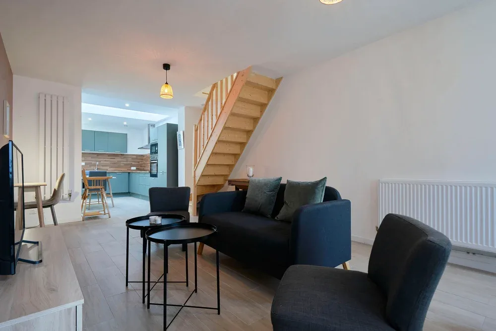 A living room with dark seating and a wooden staircase beside an open kitchen.