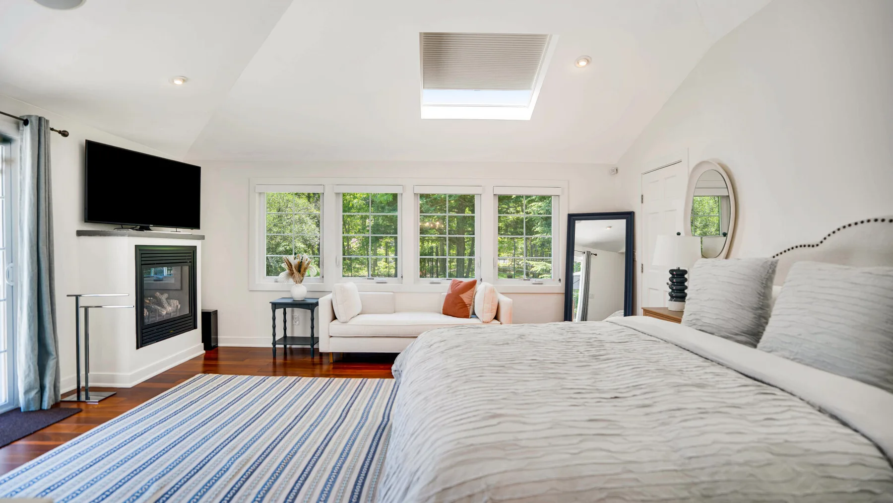 Un dormitorio luminoso con una cama grande, una alfombra a rayas y ventanas con vistas a la vegetación.