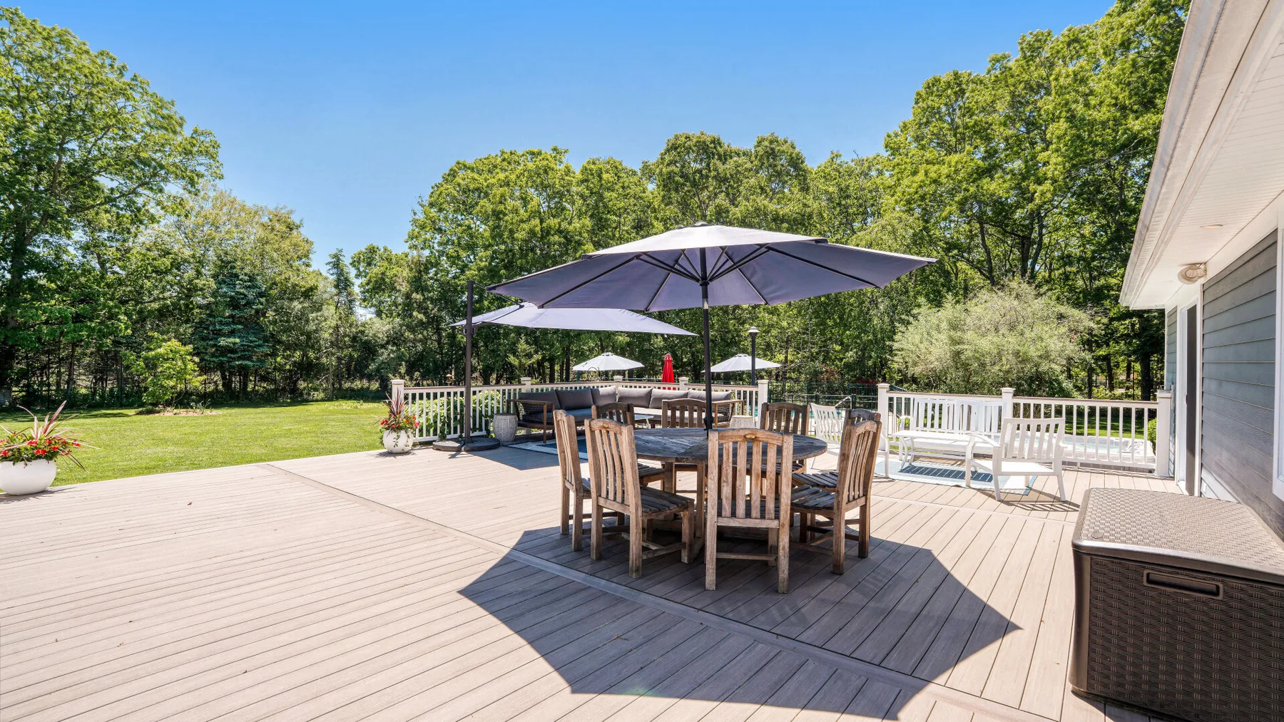 Una terraza exterior tiene una mesa de comedor bajo una sombrilla grande, con árboles al fondo.